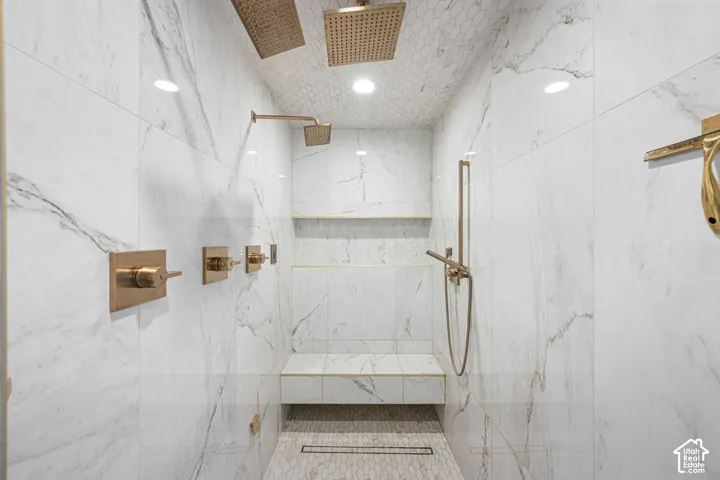 Walk-in shower featuring floor-to-ceiling large format tile, a built-in tiled bench, dual overhead rainfall showerheads, metallic-finish wall-mounted hardware, and a linear floor drain