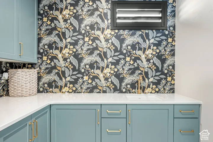 Blue cabinetry with brass-finish pulls, quartz countertop, inset sink, wall-mounted faucet, and patterned wallpaper