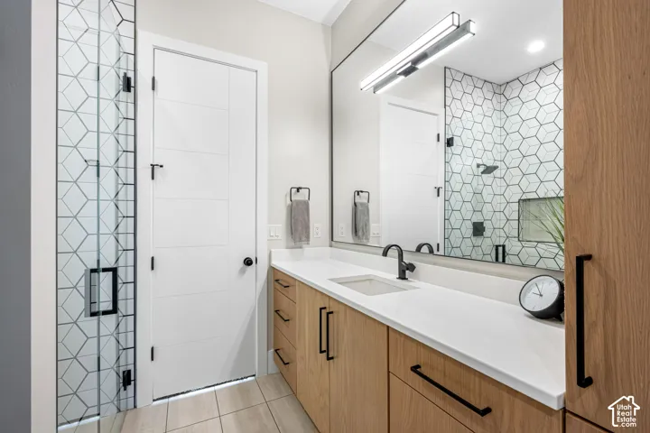 Bathroom featuring wood-finish cabinetry, white countertop with an integrated rectangular sink, matte black hardware, floor-to-ceiling glass-enclosed shower with geometric tile surround, and expansive wall-mounted mirror