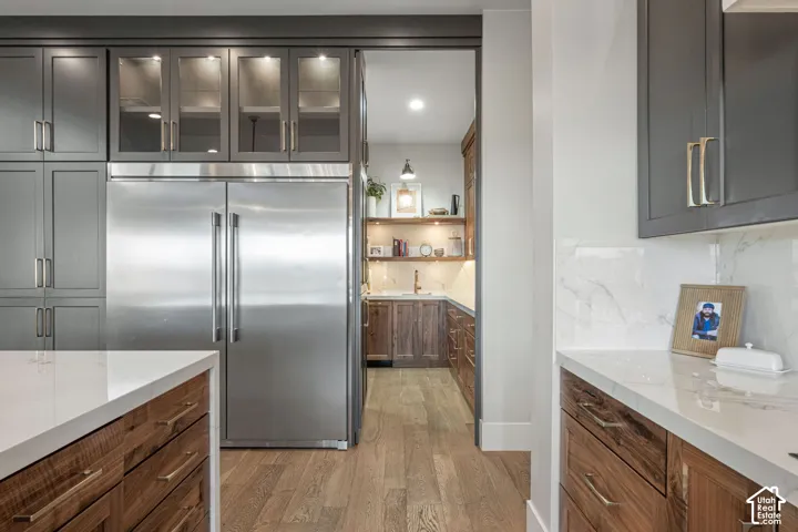 Kitchen featuring dark grey custom cabinetry, stainless steel double-door refrigerator, stone-composite countertops with marble-style veining, wood-finish flooring, and natural wood cabinetry with metal hardware