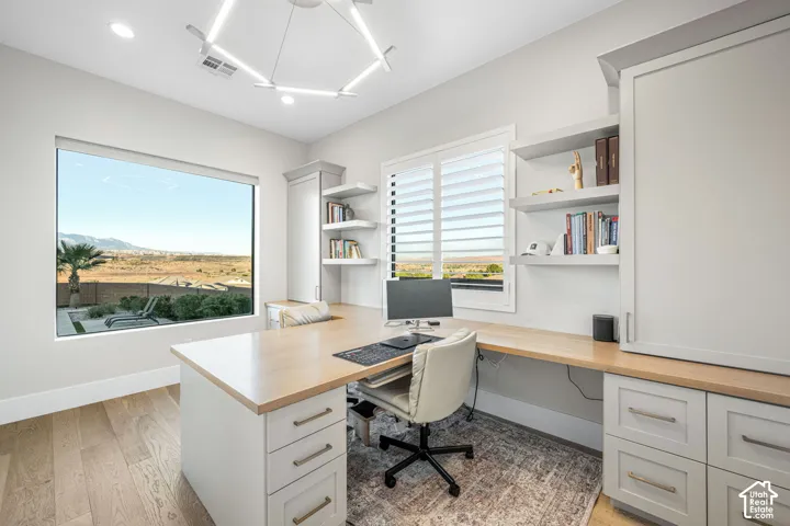 Custom built-in cabinetry with light-toned wood desktop, wood-finish flooring, plantation shutters, geometric chandelier, and large-format picture window