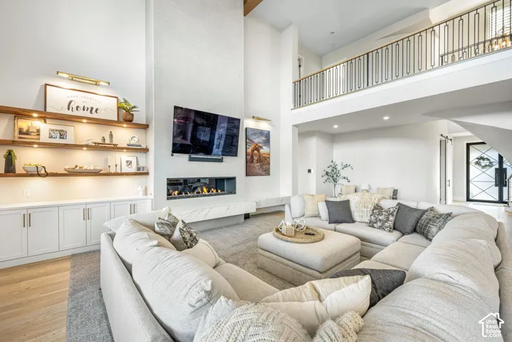 Double-height ceiling, linear fireplace with stone-finish hearth, built-in cabinetry, wood-finish floating shelving, and metal-railing mezzanine