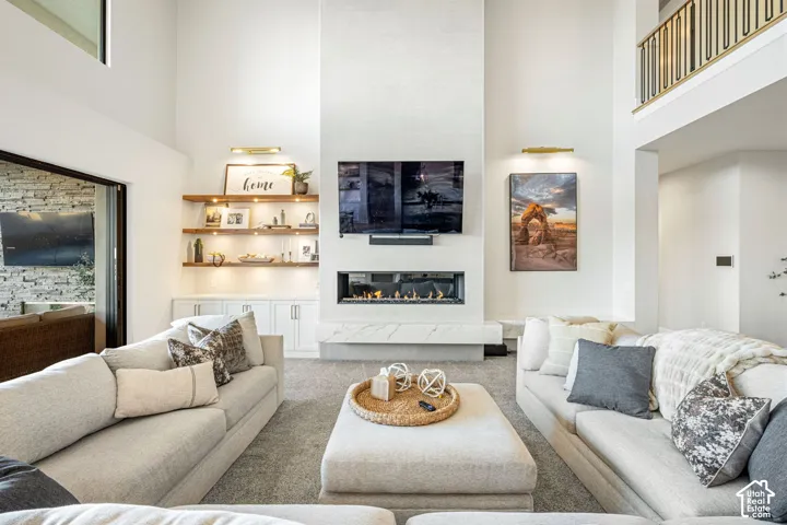 Double-height ceilings, floor-to-ceiling stone-surround fireplace with a linear insert, stone hearth, built-in cabinetry with integrated shelving, and wall-mounted brass accent lighting