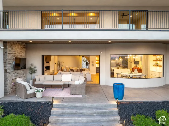 Covered concrete patio featuring a stacked stone fireplace, wide-span glass sliding door, curved glass window wall, and second-story balcony with a metal railing