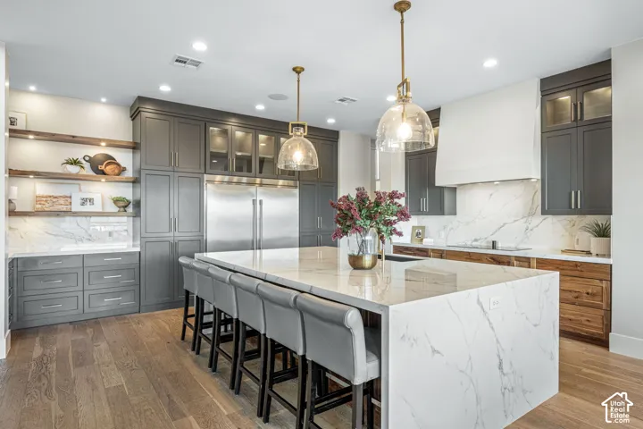 Kitchen featuring a large central island with a stone waterfall countertop, floor-to-ceiling cabinetry, custom range hood, wood-finish flooring, and open shelving with integrated lighting