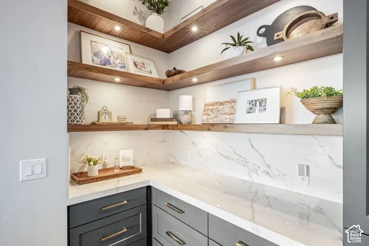 Built-in cabinetry featuring dark gray drawers with metallic pulls, white stone-finish countertop, full-height stone-finish backsplash, and three-tier recessed wooden shelving with integrated lighting