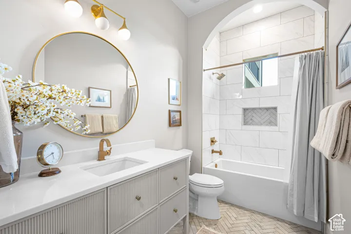 Bathroom featuring a vanity with vertical-ribbed cabinetry and a stone-finish countertop, a circular gold-frame mirror, an arched shower alcove with tile surround, and herringbone-patterned floor tile