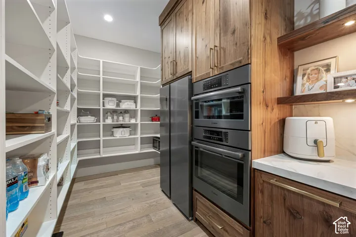 Walk-in pantry featuring white custom built-in shelving, wood-finish flooring, integrated stainless steel appliances, natural wood cabinetry, and stone-finish countertops with under-cabinet lighting