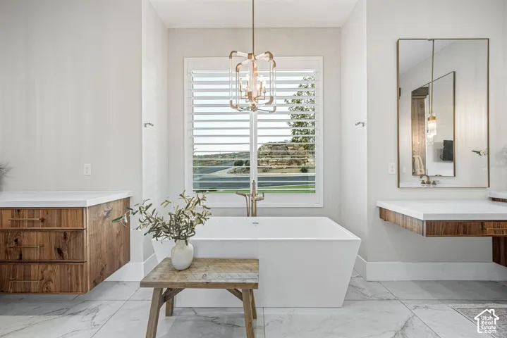 Freestanding rectangular soaking tub with wall-mounted metallic fixtures, floating wood-finish cabinetry, white solid-surface countertops, large-format tile flooring, and a geometric metal pendant light