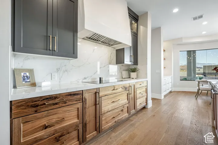 Kitchen featuring two-tone cabinetry with wood-finish lower drawers, charcoal upper cabinets, integrated range hood, stone-finish backsplash, and wood-finish flooring