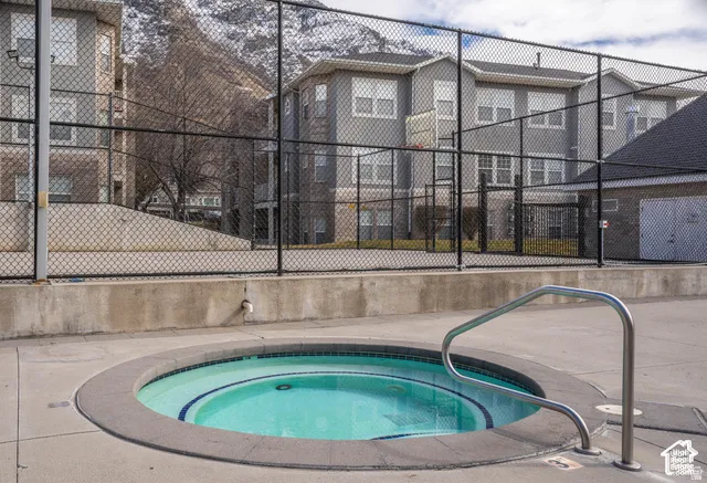 In-ground circular spa featuring a concrete surround, metal handrail, and tiled perimeter edge, set within a fenced outdoor recreation area including a paved sport court and mountain backdrop