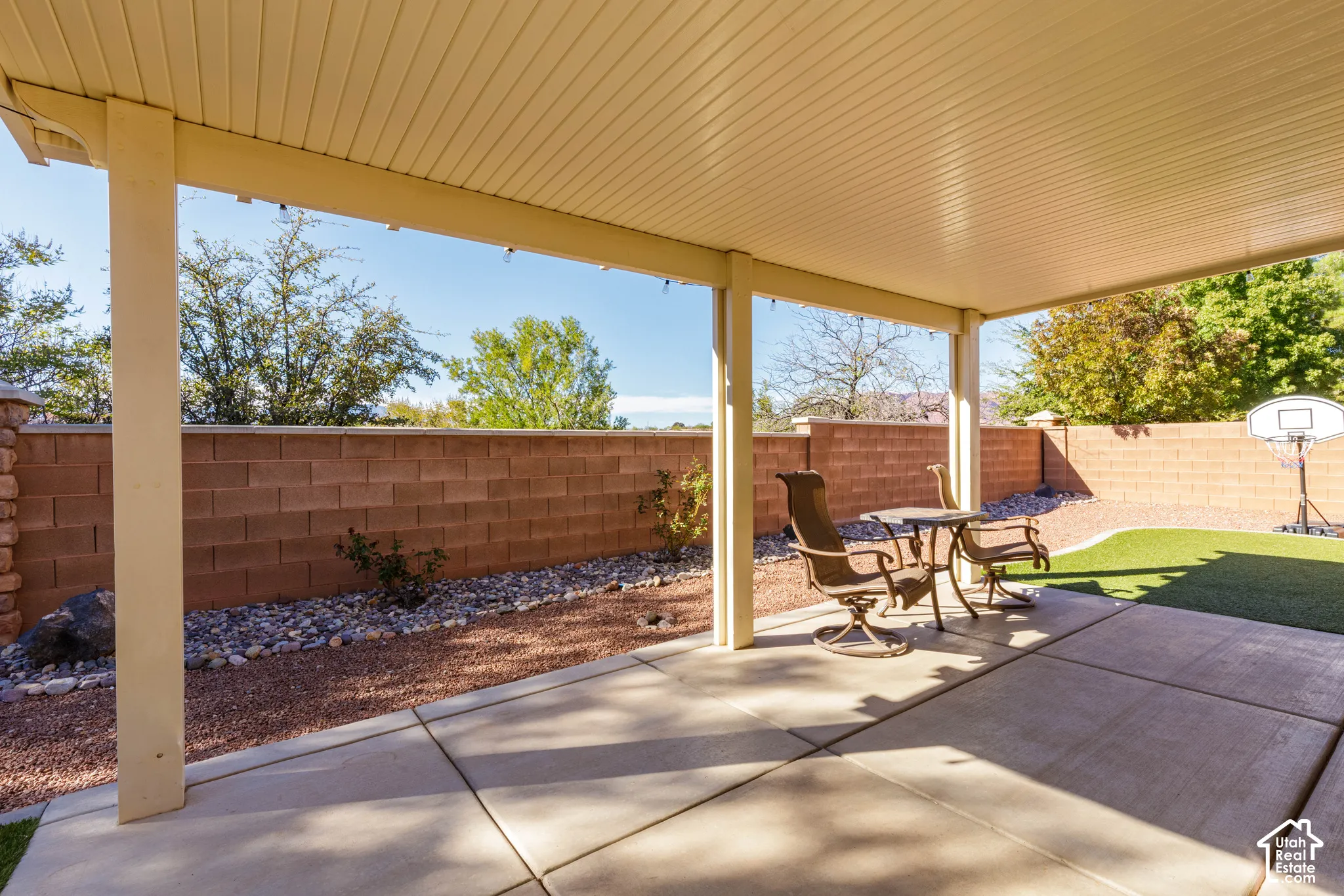 Covered concrete patio featuring a structural support beam and a perimeter masonry block wall enclosing a low-maintenance yard with stone ground cover and artificial turf