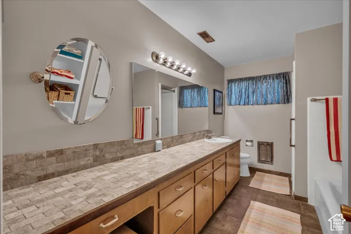 Bathroom featuring an extended vanity with tile-surfaced countertop, integrated backsplash, built-in recessed shelving, wall-mounted oval mirror, and multi-bulb vanity lighting