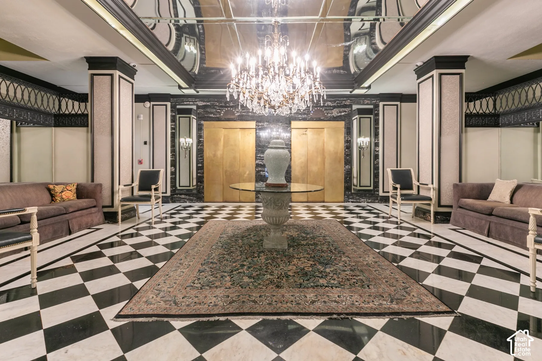 Grand lobby featuring a black and white checkerboard stone floor, dual gold-tone elevator doors, recessed ceiling mirrors, ornate crown molding, and a central tiered chandelier