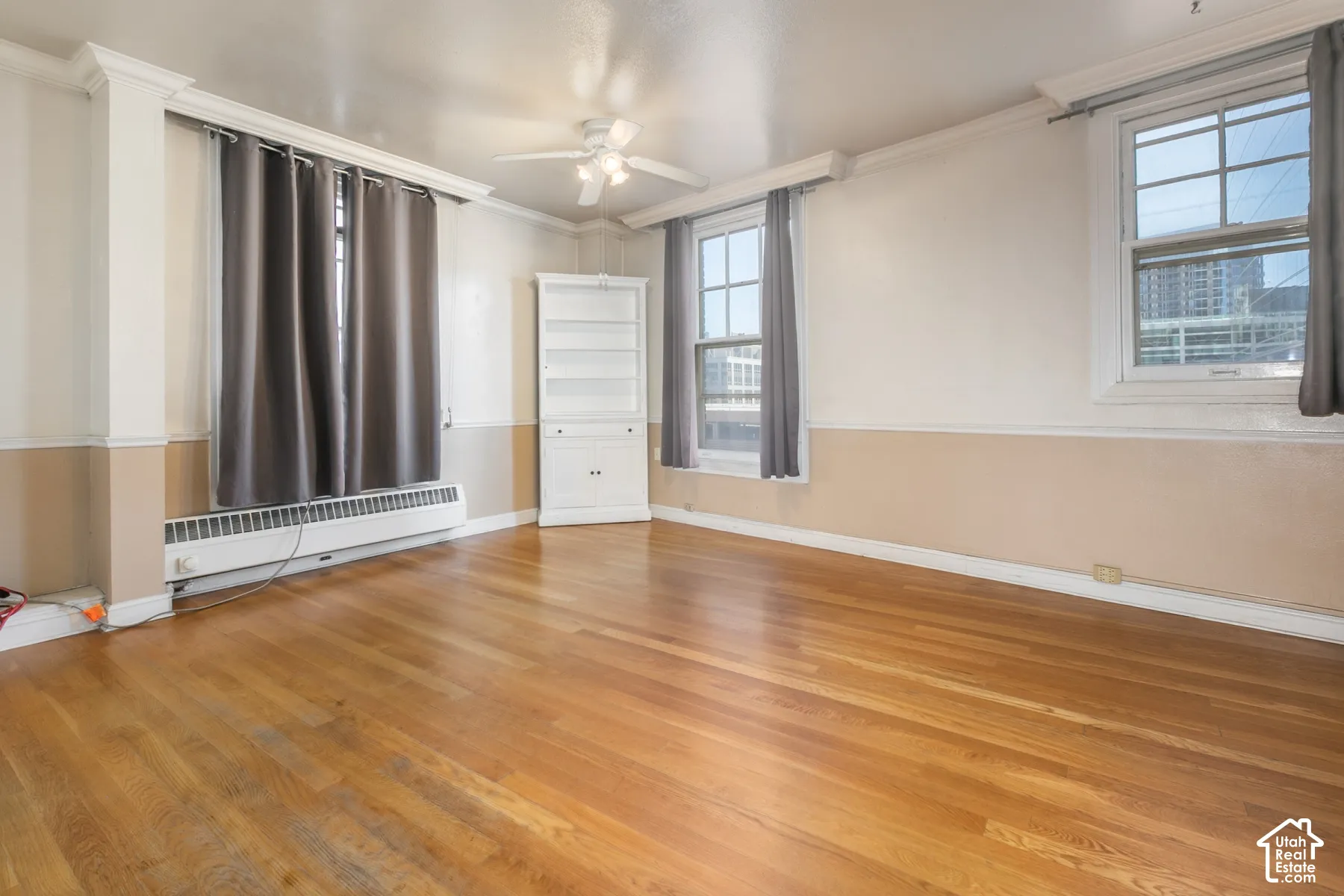 Wood-finish flooring, two-tone walls with crown molding, double-hung windows, built-in corner cabinet, and ceiling fan with integrated lighting