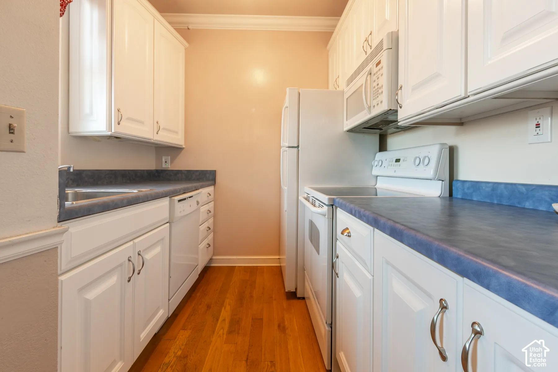 Galley kitchen featuring white raised-panel cabinetry, contrasting solid-surface countertops, wood-finish flooring, and built-in white appliances