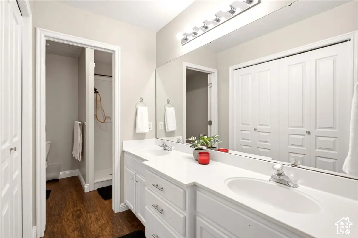 Owner suite bath, dual-sink vanity with white cabinetry, light-toned countertop, and full-width vanity mirror, featuring wood-finish flooring and bi-fold closet doors