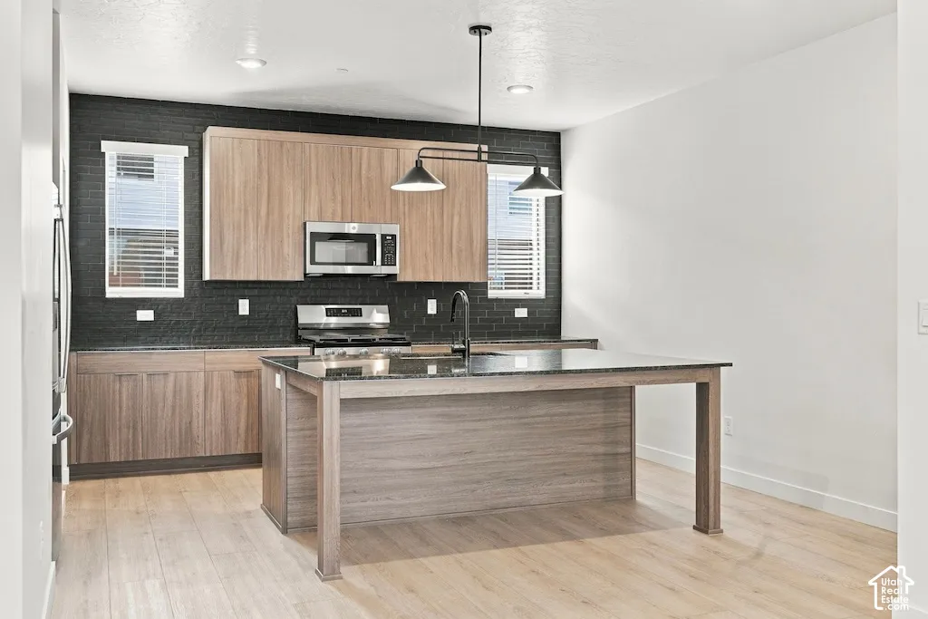 Kitchen featuring wood-finish cabinetry, a central island with a stone-top surface, dark-toned textured tile backsplash, stainless steel appliances, and multi-light pendant fixture