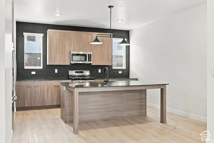 Kitchen featuring wood-finish cabinetry, a central island with a stone-top surface, dark-toned textured tile backsplash, stainless steel appliances, and multi-light pendant fixture