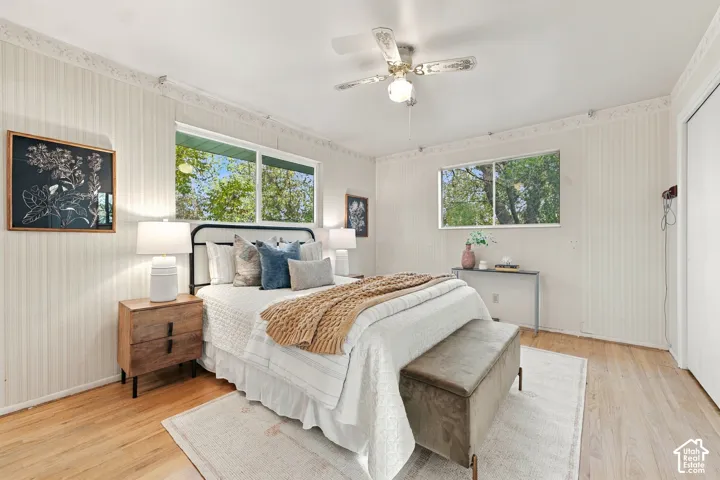 Interior room featuring vertical textured wallcoverings, light-toned wood-finish flooring, dual windows with natural greenery, decorative crown molding, and a central ceiling fan with an integrated light fixture