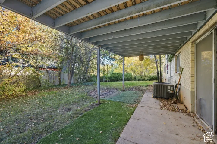 Covered lower-level concrete patio with exposed timber joist ceiling, brick exterior wall, sliding glass door, and expansive backyard
