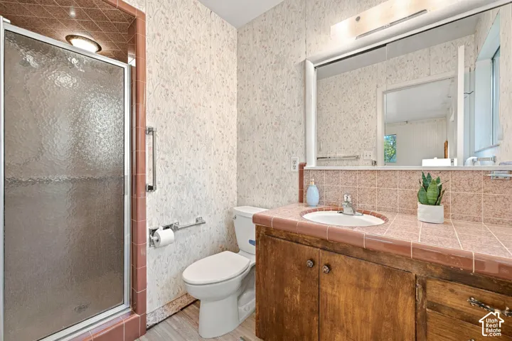 Bathroom featuring a tiled vanity countertop with an integrated oval sink, wood-finish cabinetry, a shower stall with a textured glass door, wall-to-wall mirror, and patterned wall covering