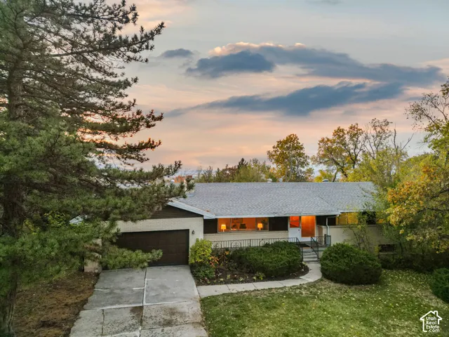 Ranch-style exterior featuring a light-toned brick facade, an attached garage, and a concrete driveway set among mature trees