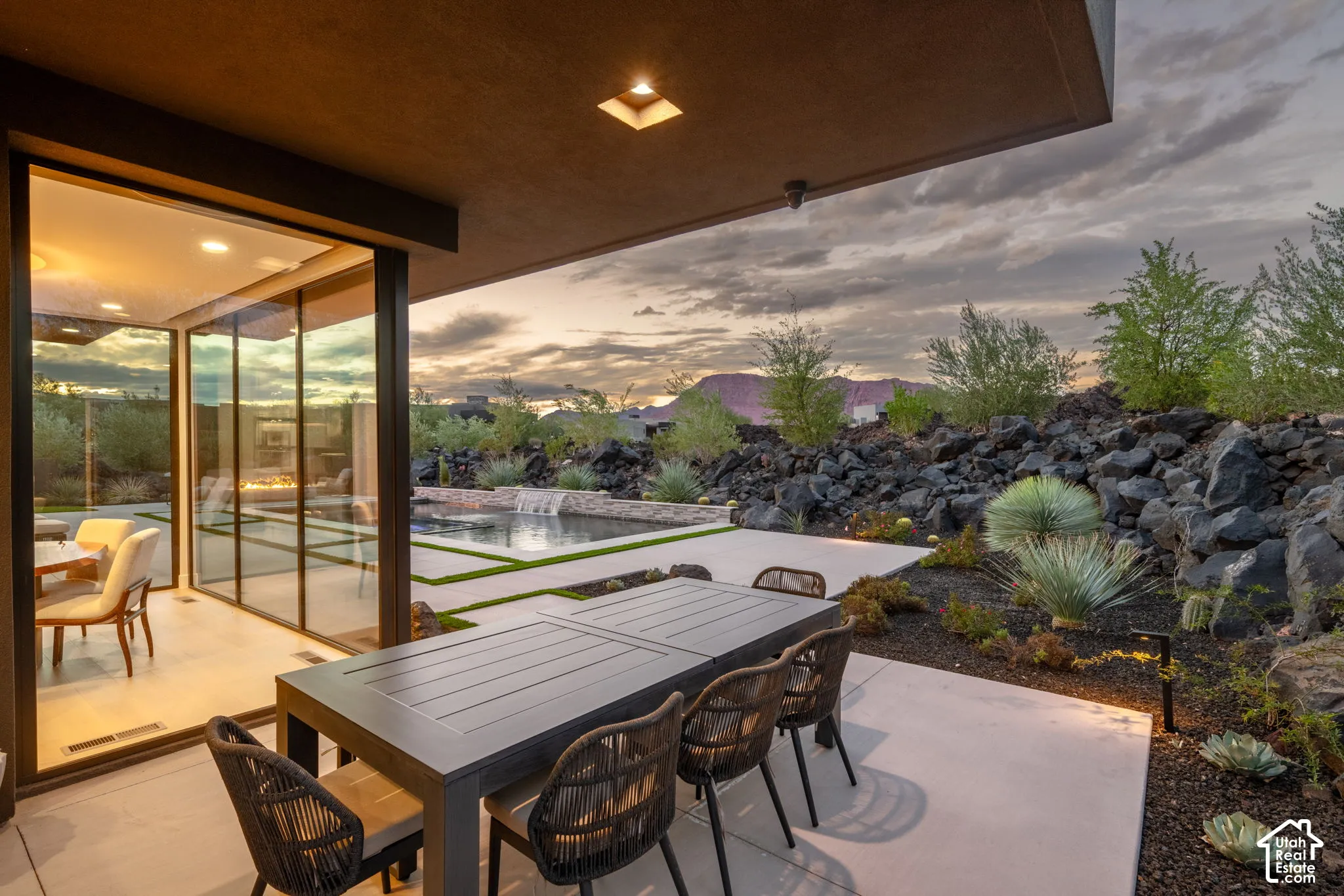 Covered concrete patio featuring floor-to-ceiling glass wall panels, an inground pool with water feature, and desert-inspired landscape with natural rock formations