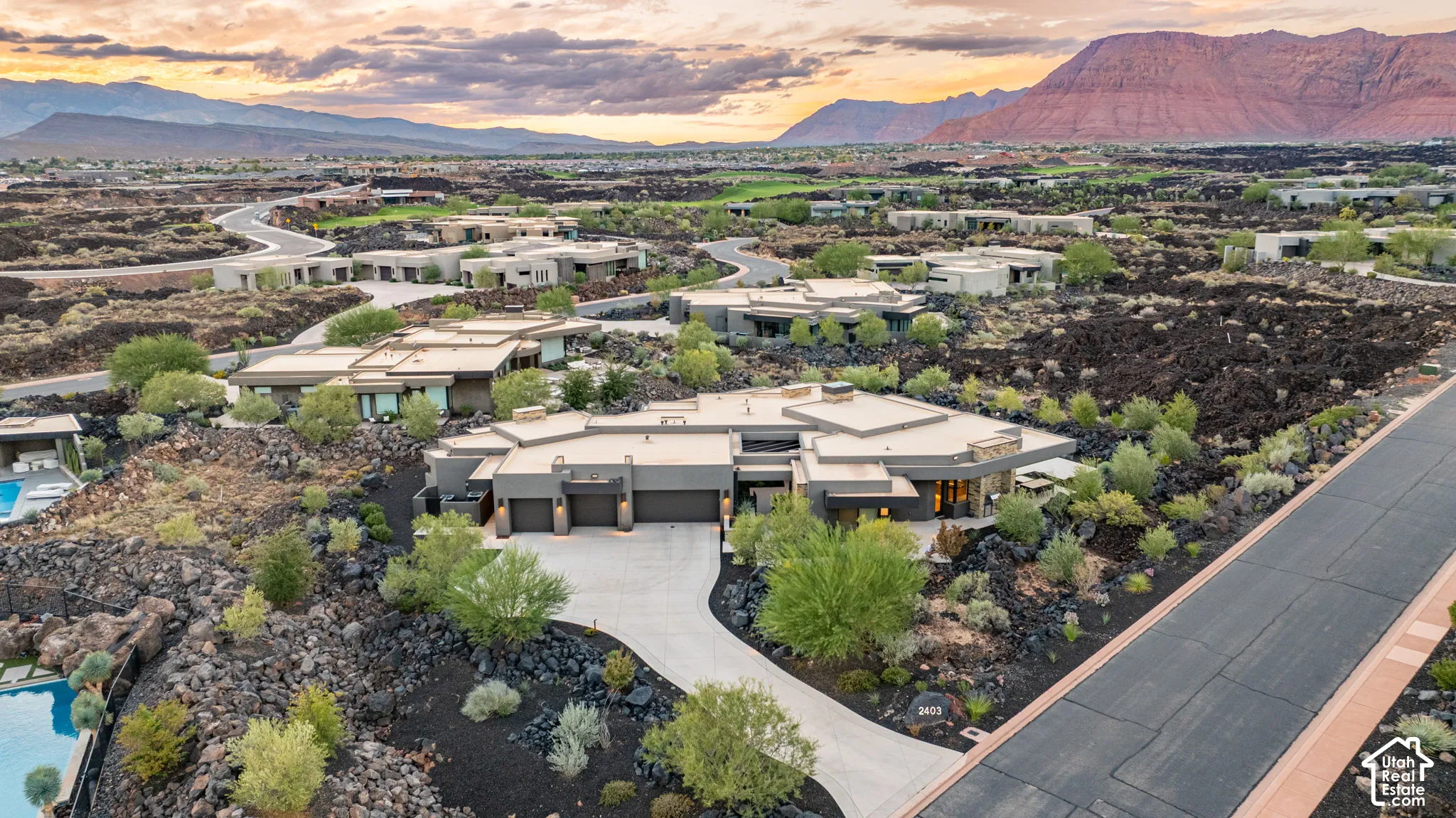 Aerial perspective of a multi-level contemporary residence featuring a three-car garage, concrete driveway, and expansive roofline, set amidst volcanic rock landscaping and a mountainous backdrop