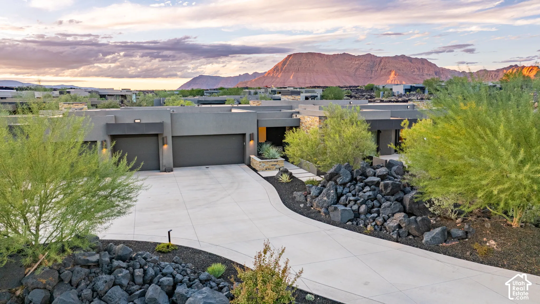 Contemporary exterior featuring a flat roofline, integrated multi-car garage, stone veneer accents, concrete driveway, and xeriscaped grounds with volcanic rock formations, framed by a prominent mountain backdrop