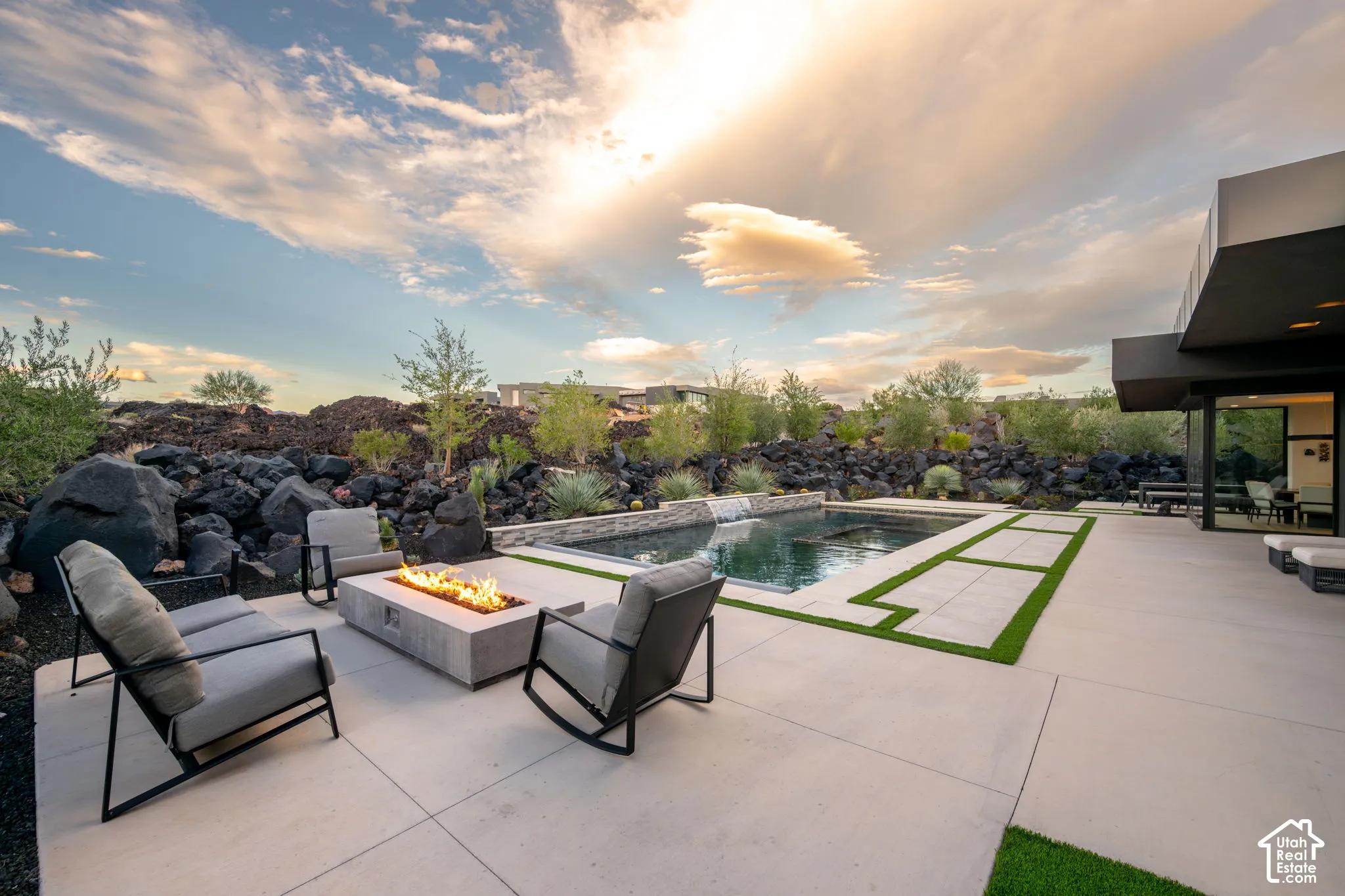 Concrete patio featuring an in-ground swimming pool with a stone water feature, built-in rectangular fire pit, and inlaid synthetic turf accents