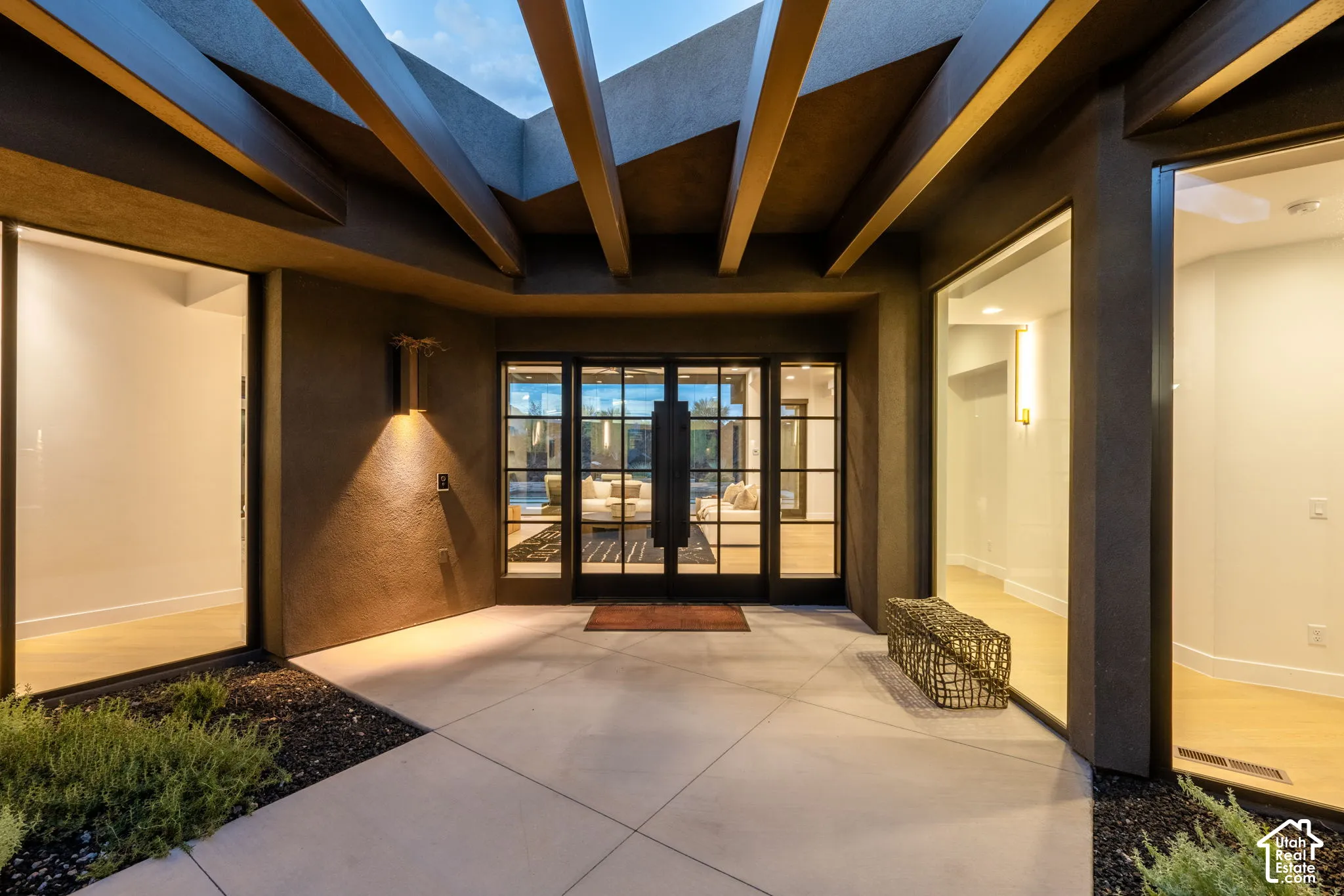 Entryway featuring a steel-framed glass double door, overhead exposed structural beams, smooth concrete patio, and large-format glass window panels