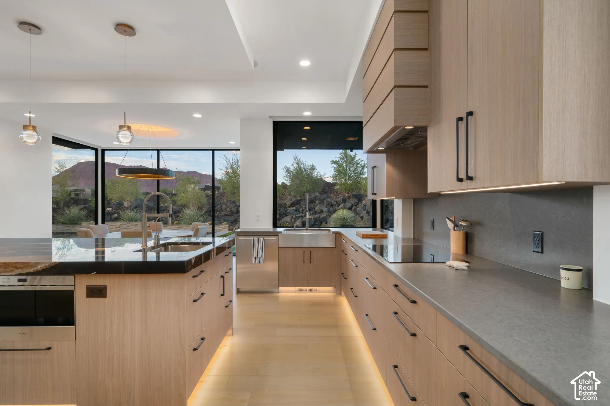 Contemporary kitchen featuring light wood-finish cabinetry, matte stone countertops, integrated stainless steel appliances, a farmhouse-style sink, and floor-to-ceiling windows