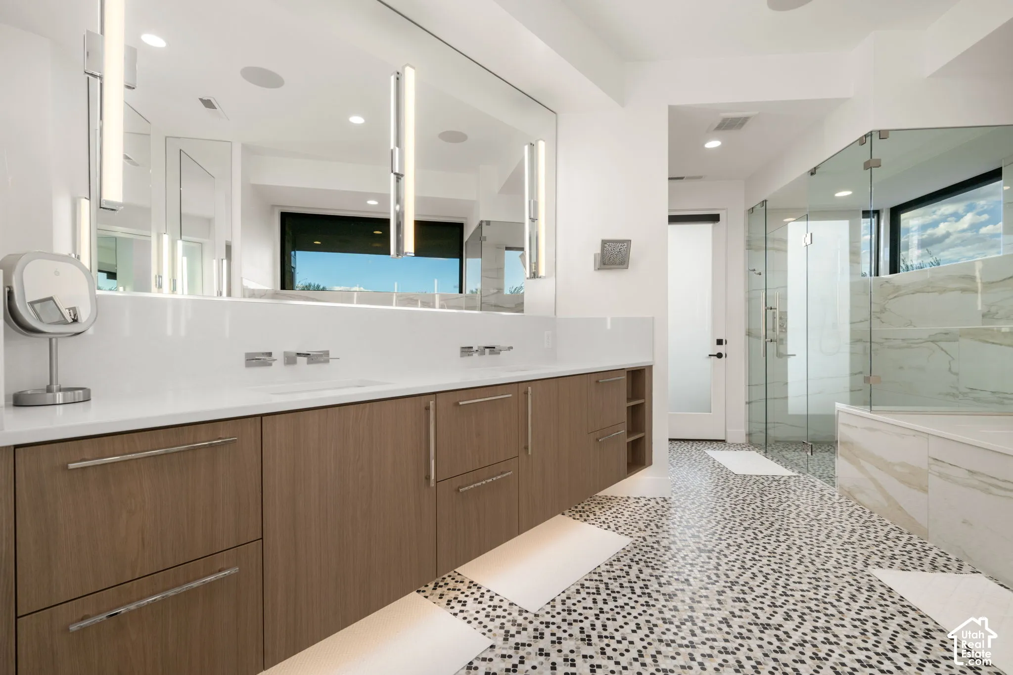 Dual vanity with wood-finish cabinetry, white countertop, integrated sinks, wall-mounted fixtures, and a glass-enclosed shower with stone-finish surround