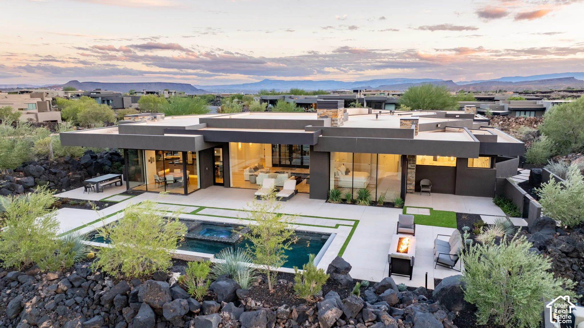 Modern single-story exterior featuring expansive floor-to-ceiling glass walls, integrated concrete patio, negative-edge swimming pool with built-in spa, and stone-accented chimneys