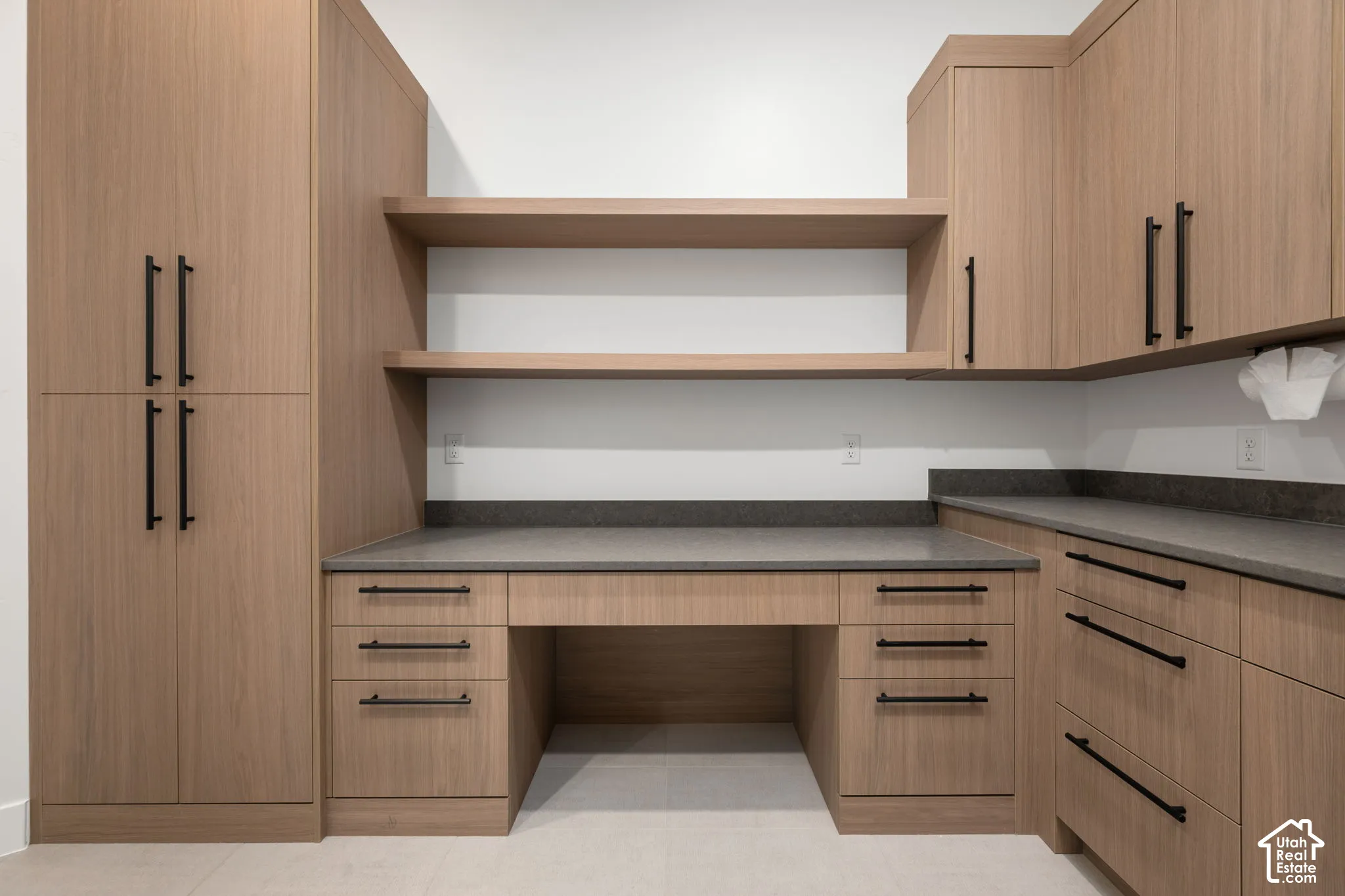 Custom wood-finish cabinetry featuring matte black hardware, integrated desk workspace, dual floating shelves, and dark solid-surface countertops