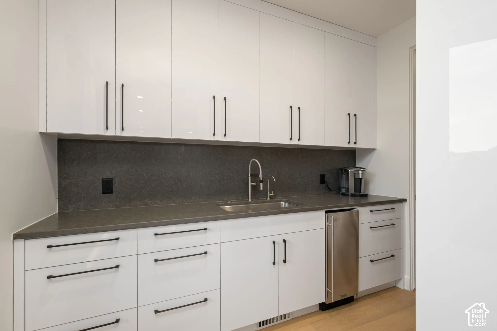 High-gloss white cabinetry with matte black hardware, dark solid-surface countertops, full-height backsplash, undermount sink with gooseneck faucet, and stainless steel under-counter beverage chiller