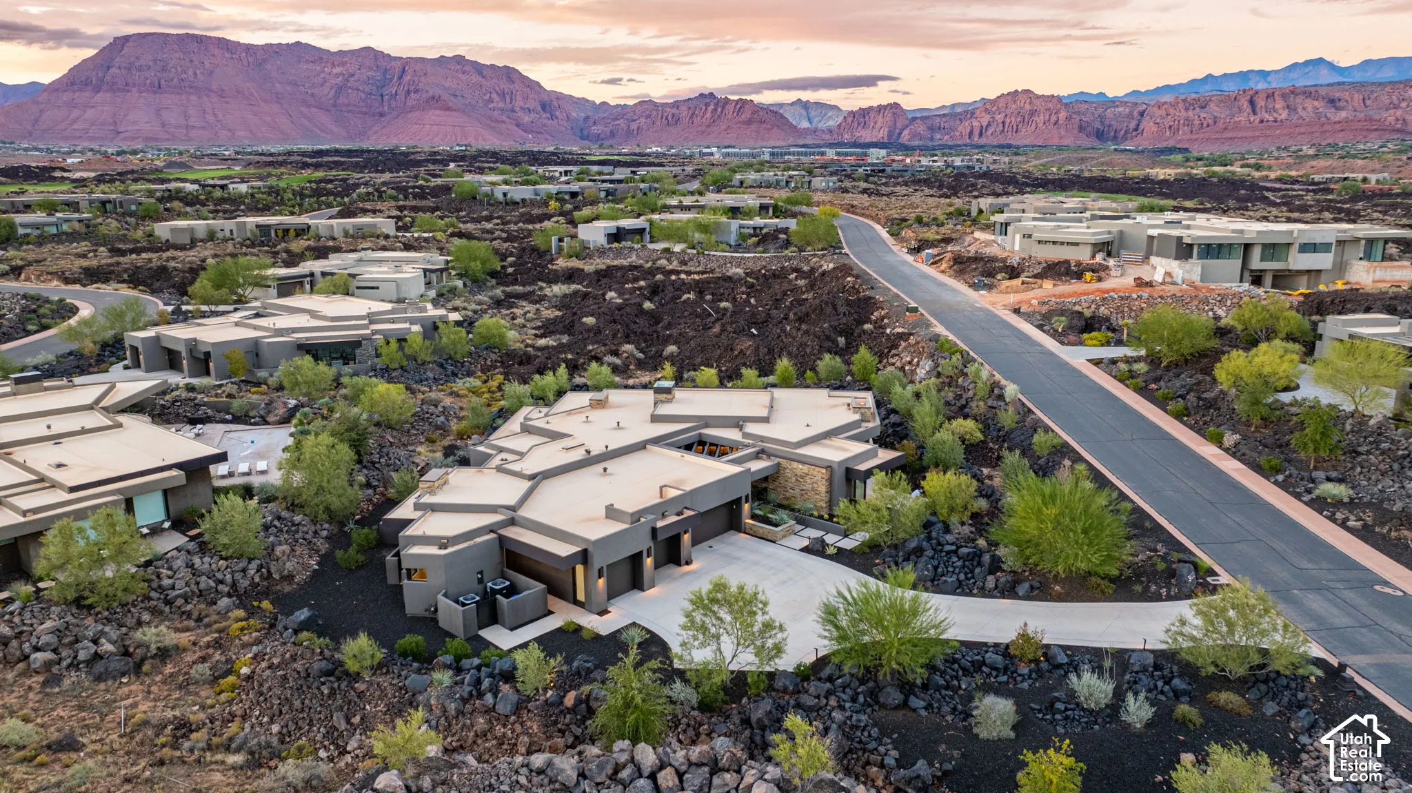 Aerial perspective of a contemporary residence featuring a multi-faceted flat roofline, stone veneer accents, integrated multi-car garage, and concrete driveway set against a backdrop of red rock mountain formations