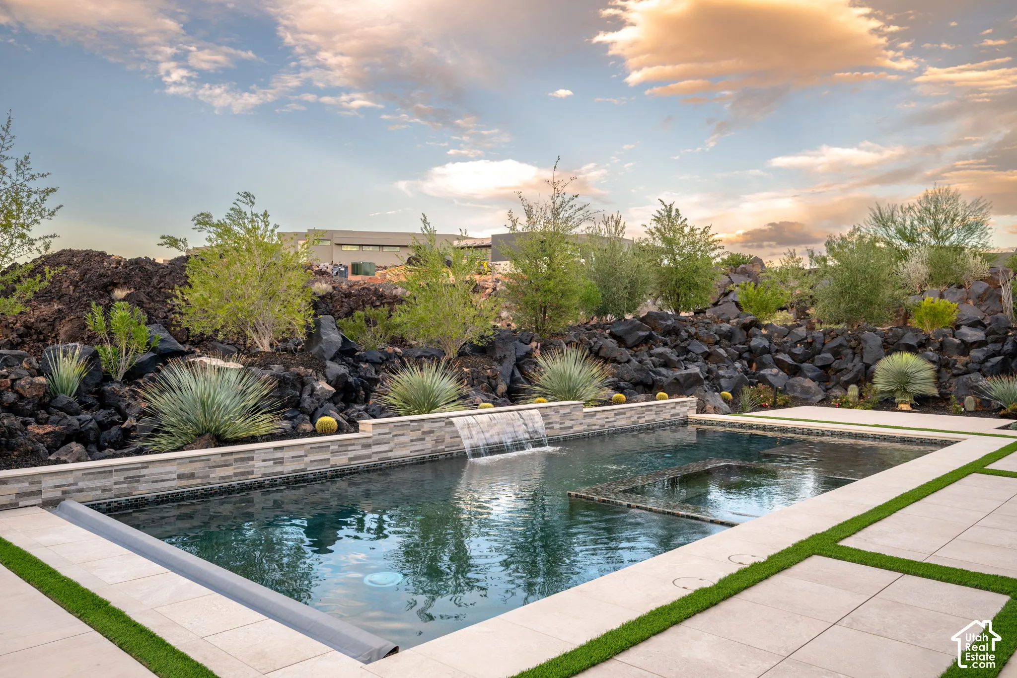 Custom swimming pool featuring a stone-clad water feature, integrated spa, and rectangular concrete paver decking with turf accents, set against a natural volcanic rock landscape