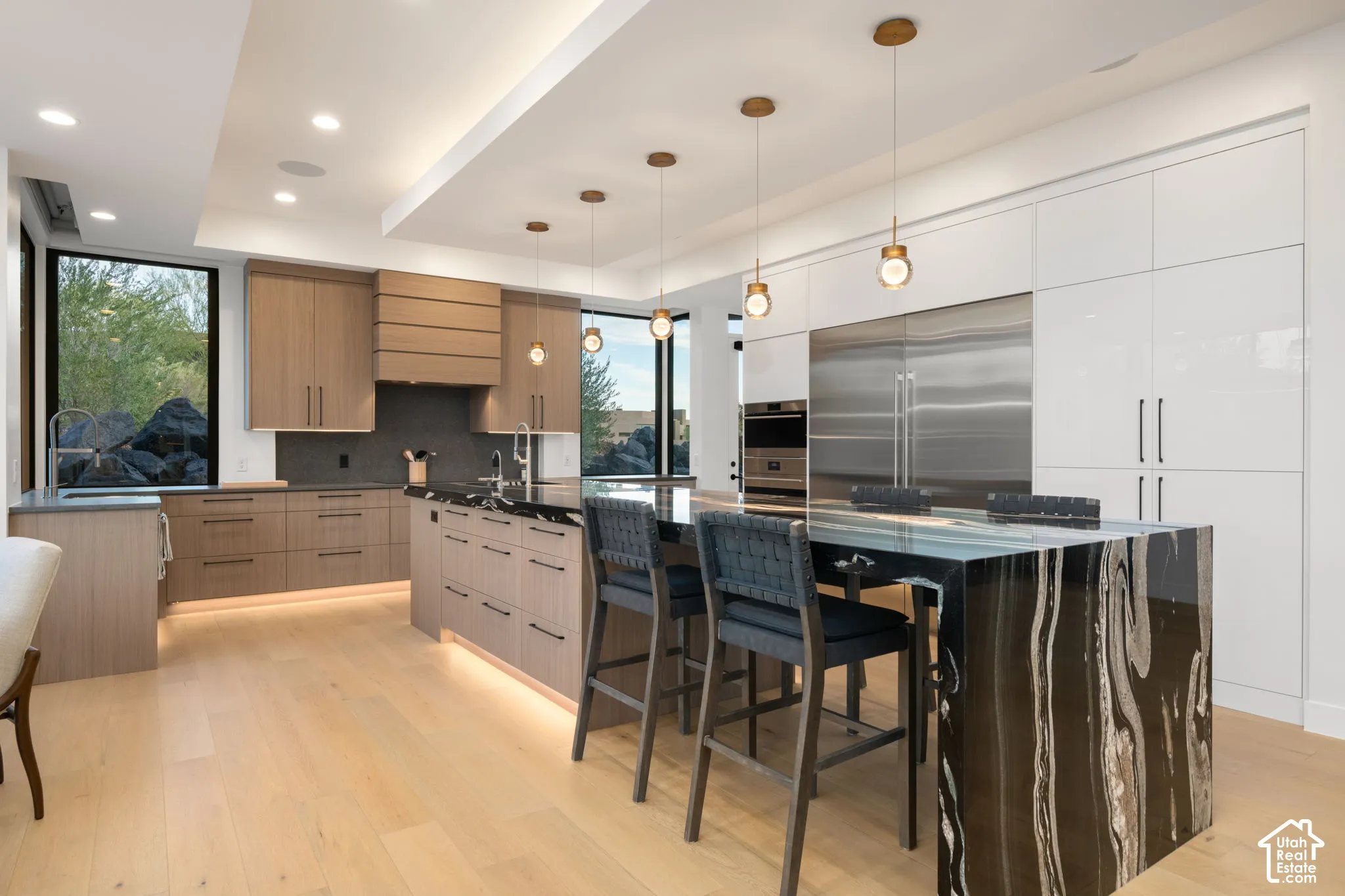 Kitchen featuring wood-finish cabinetry, a waterfall-edge stone island with seating, integrated stainless steel appliances, tiered ceiling with recessed lighting, and floor-to-ceiling windows