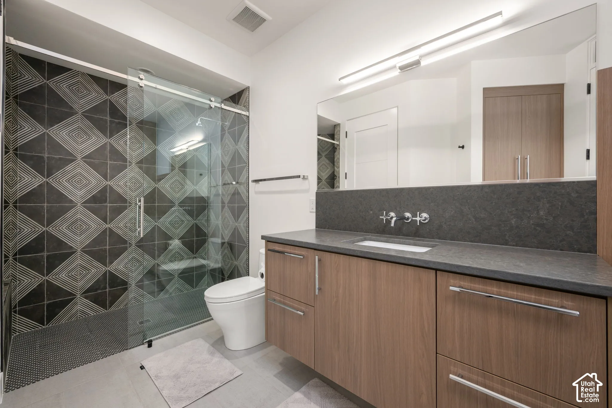 Bathroom featuring a floating wood-finish vanity, solid-surface countertop, wall-mounted faucet, walk-in glass-enclosed shower with geometric tile surround, and an integrated full-width mirror
