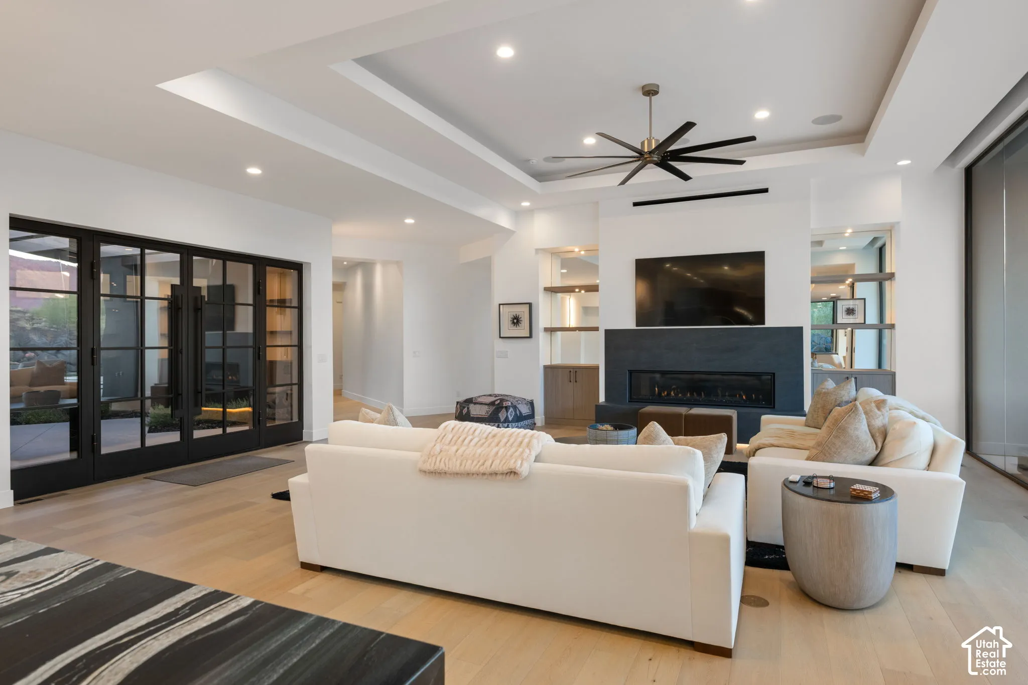 Living area featuring a tray ceiling with recessed lighting, linear fireplace with floor-to-ceiling stone surround, light-tone wood-finish flooring, built-in shelving, and multi-panel glass doors