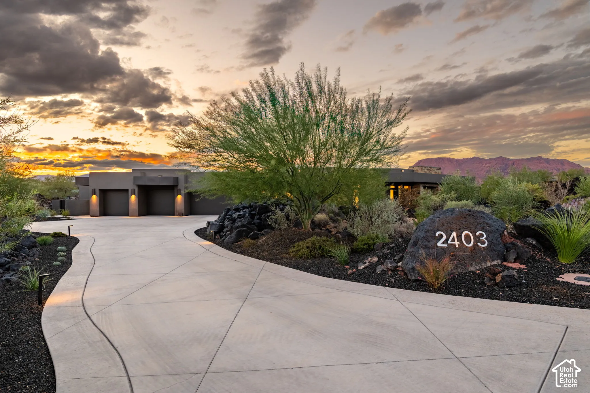 Curved concrete driveway featuring low-maintenance desert landscaping, mature native trees, natural rock formations, and a multi-car garage