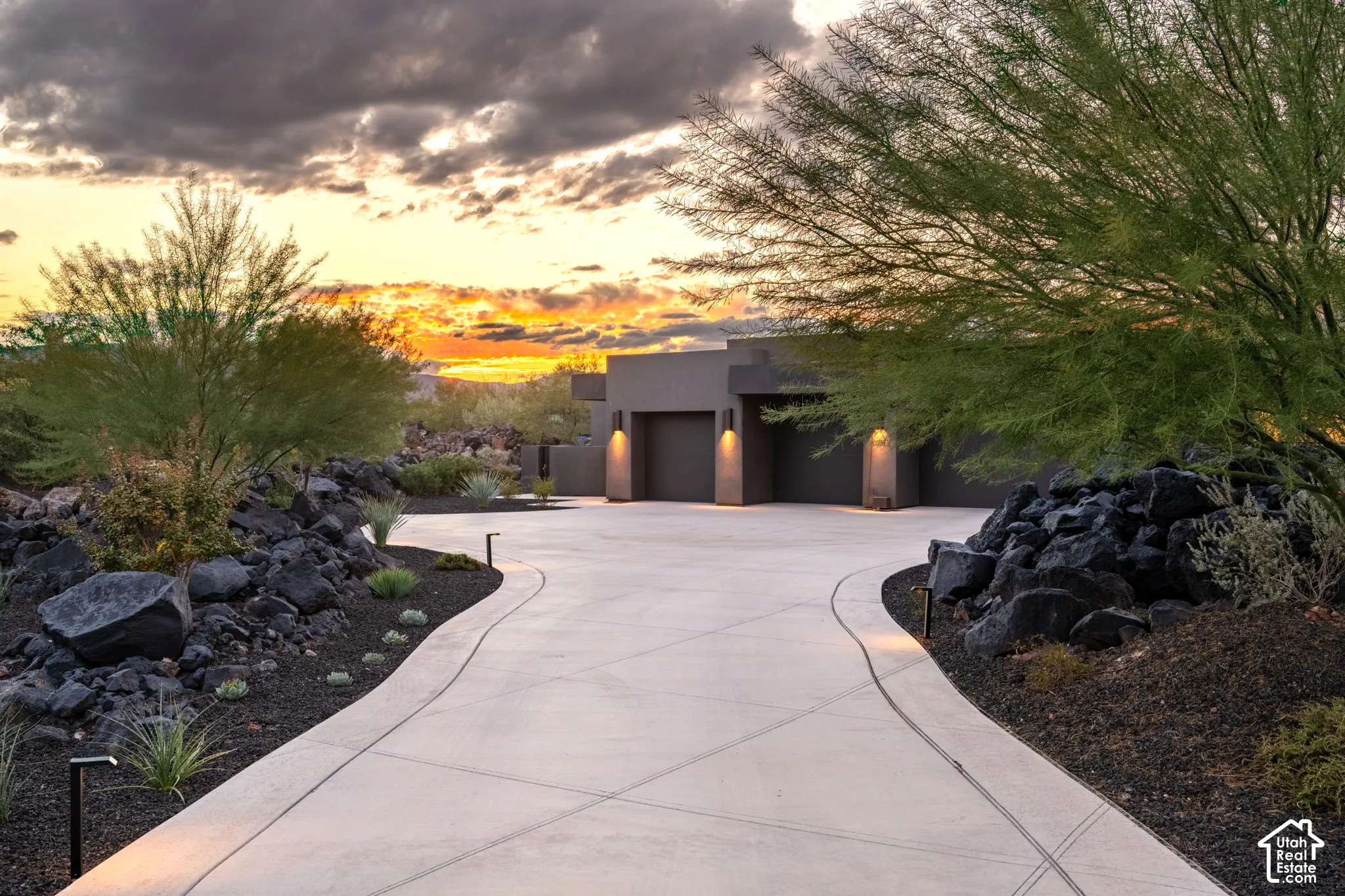 Concrete driveway featuring geometric score lines, multi-car garage, and desert landscaping with dark volcanic rock accents