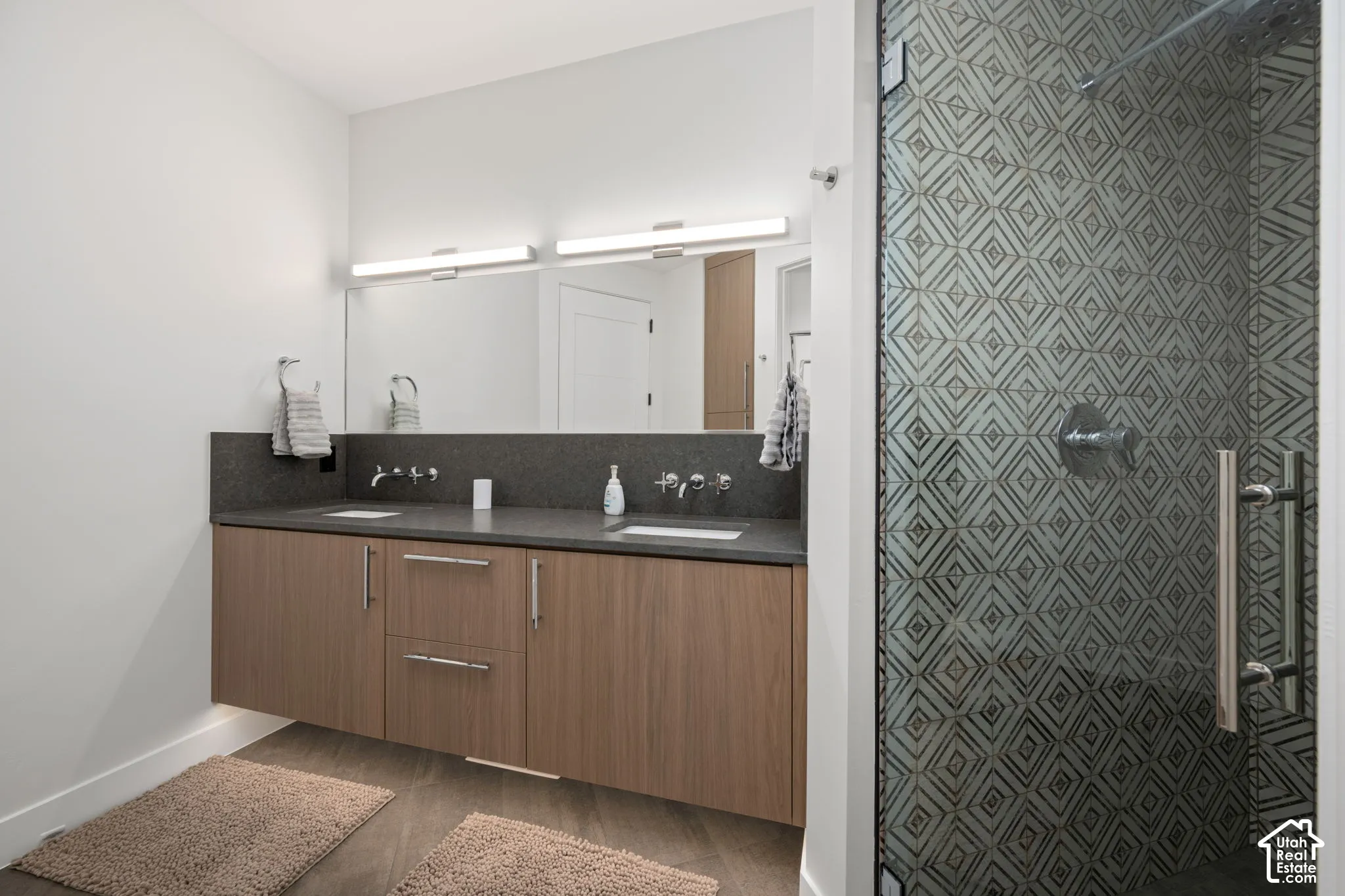 Floating vanity with wood-finish cabinetry, dual undermount sinks, quartz countertop, full-width mirror with integrated lighting, and shower enclosure featuring geometric patterned tile