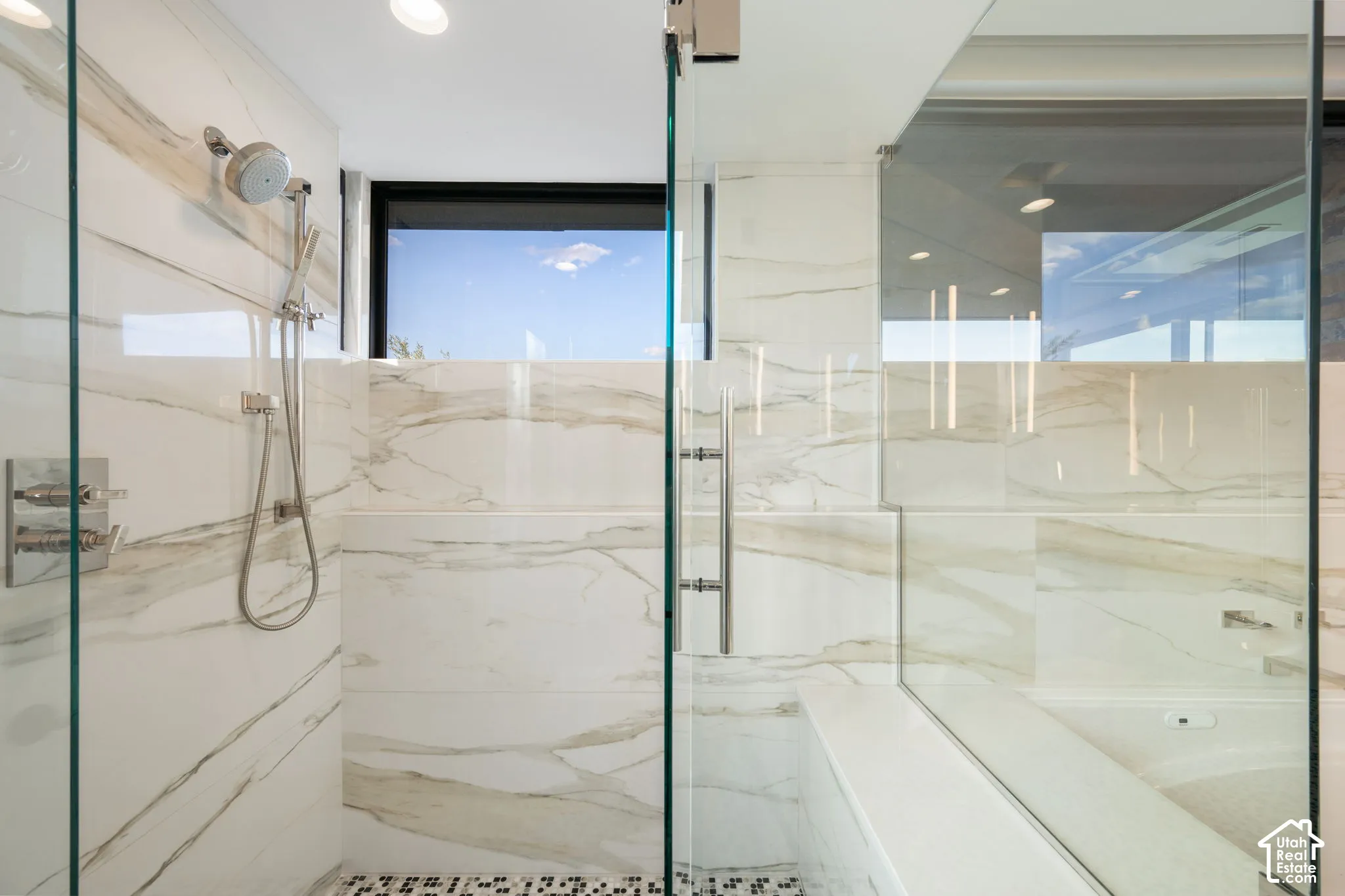 Walk-in shower featuring large-format veined tile surround, frameless glass enclosure, polished chrome rainfall shower head with handheld attachment, and geometric patterned floor tile
