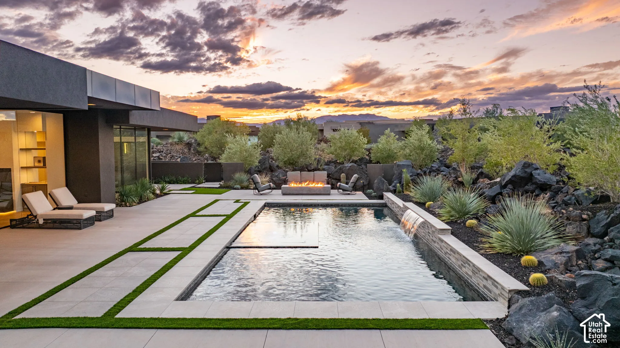 Rear exterior hardscape featuring a rectangular in-ground pool with a stone-clad water feature, integrated turf-accented concrete patio, built-in fire pit, and desert landscaping