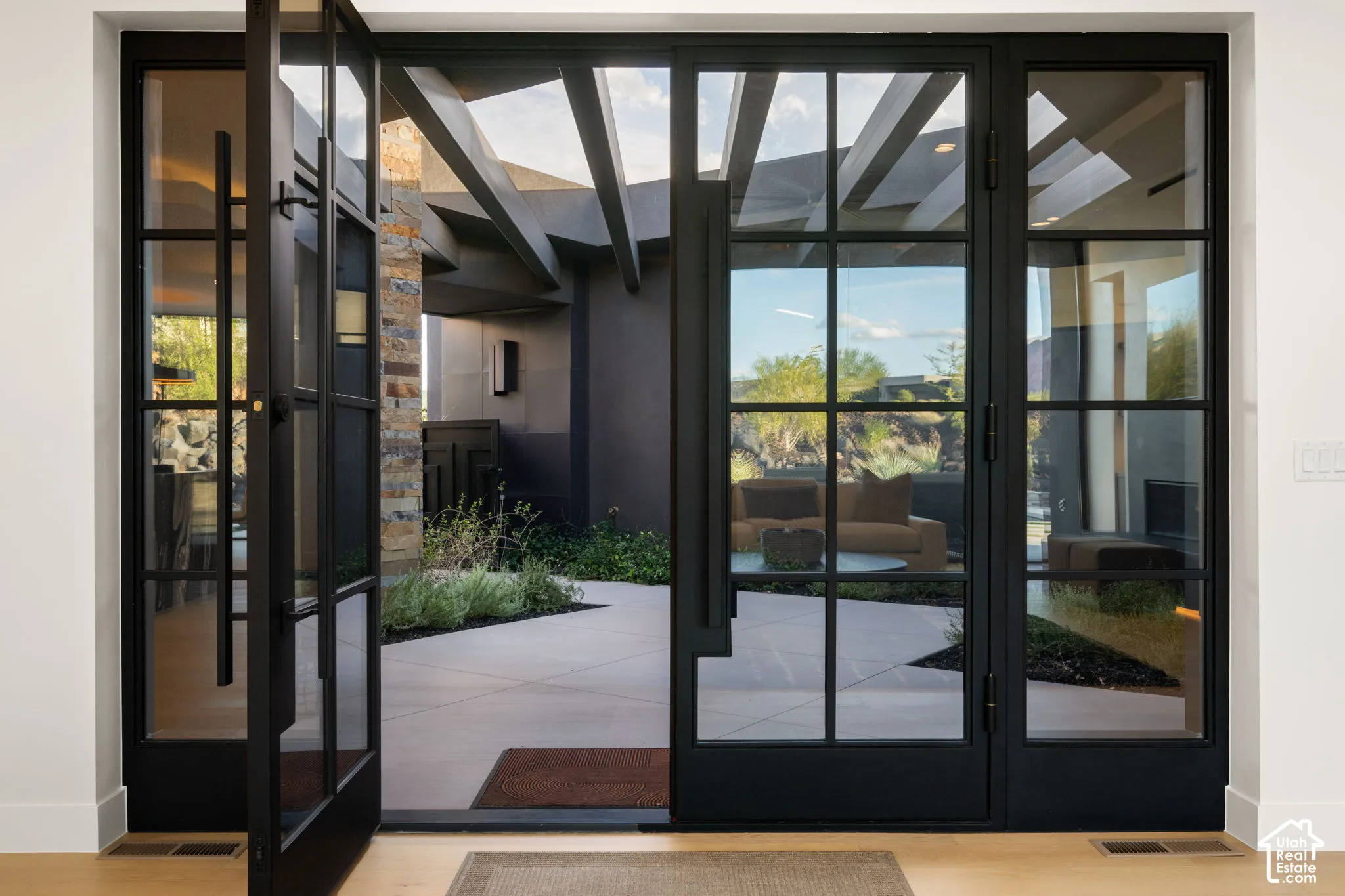 Floor-to-ceiling iron-framed glass entry doors, open-air courtyard with geometric concrete pavers, exposed structural timber beams, and natural stone masonry accent wall
