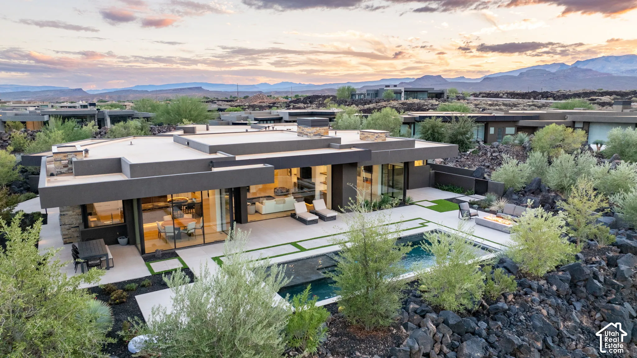 Modern architectural estate featuring an expansive flat roofline, floor-to-ceiling glass walls, integrated in-ground pool, stone exterior accents, and expansive patio space set against a mountainous horizon