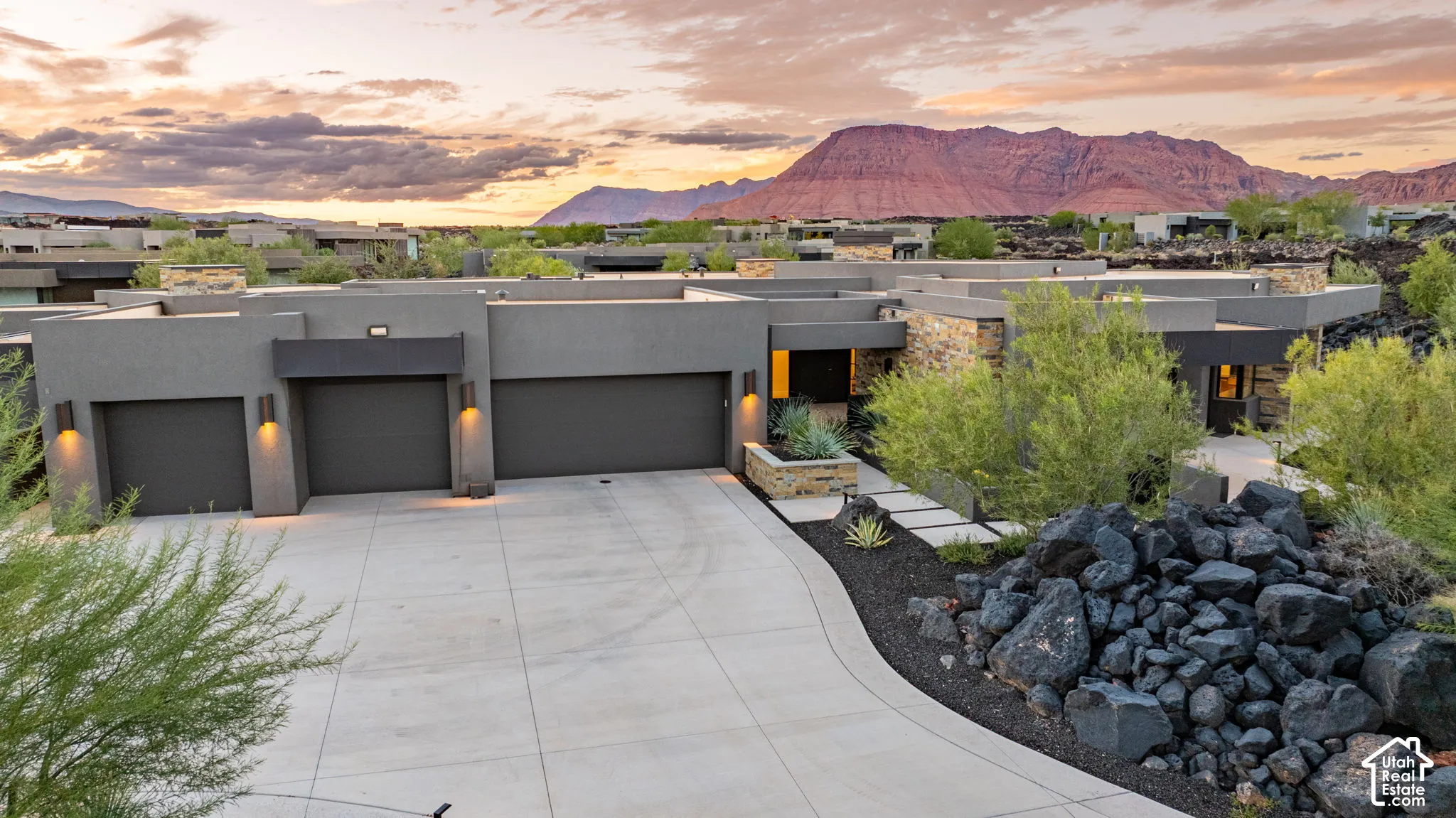 Contemporary residence featuring a multi-bay garage, textured stone facade accents, concrete driveway, and natural rock landscaping, set against a backdrop of prominent red rock mountains